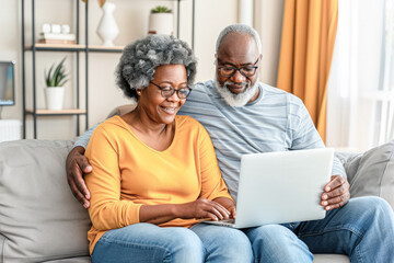Obraz premium Happy mature senior couple using laptop sit on sofa at home. Smiling senior old adult man and woman using laptop to shop online. Retirement and happy senior lifestyle concept. 