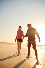 Loving Retired Senior Couple On Vacation Walking Along Beach Shoreline Holding Hands At Sunrise