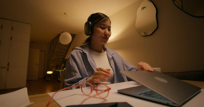A woman in casual clothes finishes a video chat on a laptop sitting at a table at home. She takes off her headphones and closes her laptop