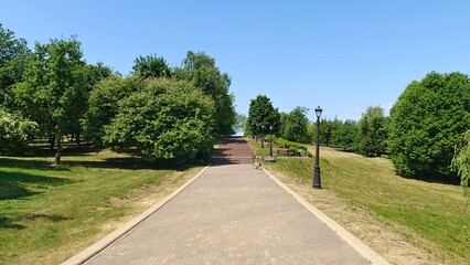 Concrete tile stairs lead from a city park to a city block. Nearby are mowed grassy lawns, various deciduous trees, metal lampposts and a bicycle. Summer sunny weather and blue sky