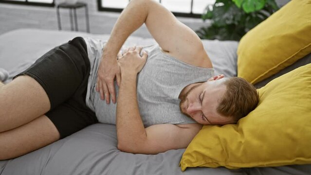 Young bearded man in pain lying on side in bedroom gripping stomach, showcasing discomfort and illness at home.