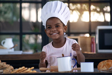 Happy little African girl wear chef hat in bakery kitchen, she learned about baking