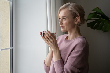 A woman stands by the window, holding a warm cup of tea in her hands. She gazes outside, lost in...