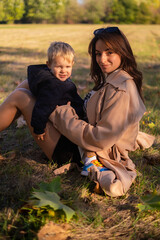 A mother and son embrace and play in the spring park. The boy holds green tree leaves while the...