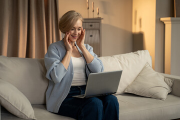 A joyful woman is seated on a sofa with a laptop on her knees, clapping her hands with an expression of elation. The warm, comfortable ambiance of the living room suggests a personal triumph
