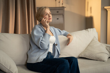 A cheerful middle-aged woman sat comfortably on the couch, holding her phone to her ear with a...