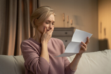 A concerned elderly woman is seated, holding a letter in her hand, with a look of distress apparent on her face. The warm lighting suggests that the scene takes place indoors, possibly during the