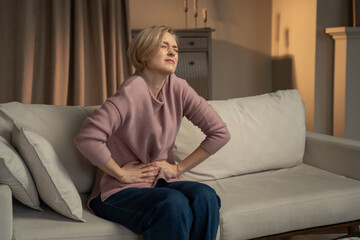 A woman is seated on her living room couch, clutching her abdomen in discomfort, her face...