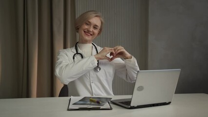 A smiling female doctor wearing a white coat and stethoscope sits at a desk with a laptop and a digital tablet, making a heart shape with her hands, likely conveying compassion and care to a patient - Powered by Adobe