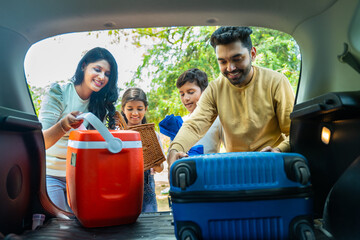 Indian couple with helping kids taking out luggage or picnic basket taking out from car boot -...