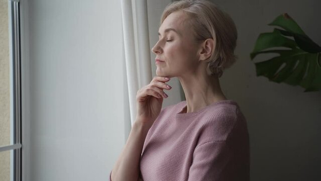 A woman with short hair, dressed in a light pink sweater, stands contemplatively near a window. Her arms are crossed as she looks out, seemingly lost in thought. The overcast light suggests it might