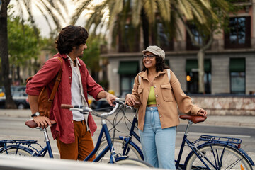 Cheerful gen z couple is standing on the city street with bicycles and chatting.