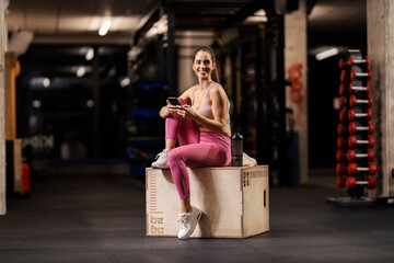 A fit sportswoman is sitting on a box in a gym and typing on her phone.