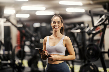 A happy female fitness instructor is standing in a gym with tablet in her hands and smiling at the camera.
