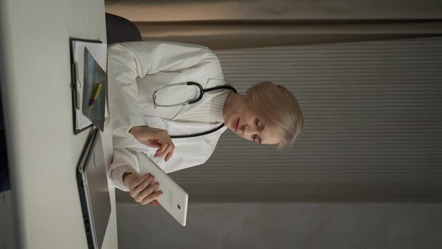 A focused female healthcare professional, wearing a white lab coat and a stethoscope, sits at a desk attentively examining digital records on a tablet. Nearby lies a closed laptop and medical