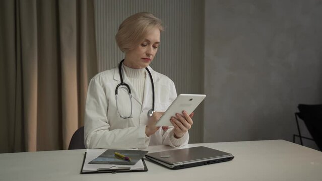 A focused female healthcare professional, wearing a white lab coat and a stethoscope, sits at a desk attentively examining digital records on a tablet. Nearby lies a closed laptop and medical