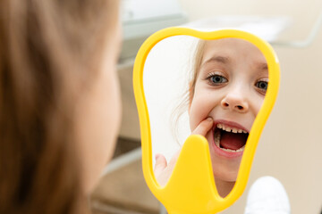 Lovely little girl looking in the mirror at her teeth after teeth surgery in a pediatric stomatology. A little girl sits in a dental chair and examines her cured teeth while looking in the mirror