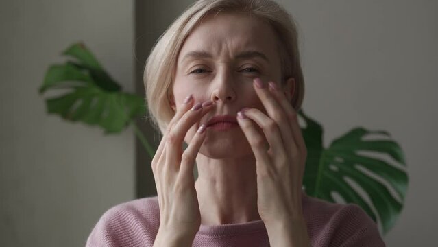 A contemplative mature woman is captured indoors while gently touching her forehead with both hands, appearing introspective or concerned. She wears a soft pink sweater, and her expression conveys