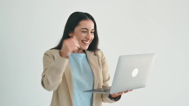 Joyful excited indian or arabian brunette business woman, dressed in elegant suit, manager, holding laptop while standing on isolated white background, rejoicing in success, gesturing with fist, smile
