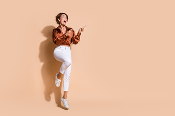 Fototapeta na wymiar Full length photo of funky impressed lady dressed brown top jumping pointing two fingers empty space isolated beige color background