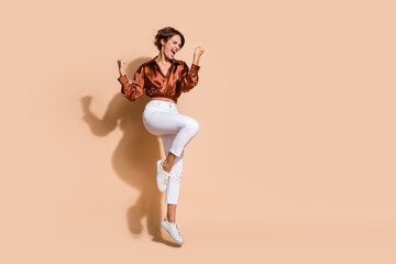 Fototapeta na wymiar Full length photo of excited lucky lady dressed brown top jumping high rising fists empty space isolated beige color background