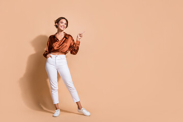 Fototapeta na wymiar Full length photo of cheerful positive lady dressed silky shirt pointing finger empty space isolated beige color background