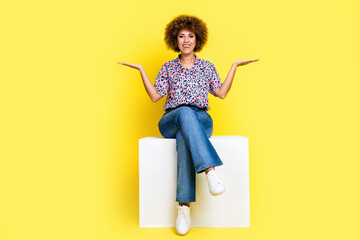 Full length photo of pretty cute woman wear print shirt sitting cube comparing arms empty space isolated yellow color background