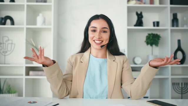 Positive successful arabian or indian woman with headset, female teacher, business consultant sitting in the office during online consultation with client, smiling, greeting interlocutor
