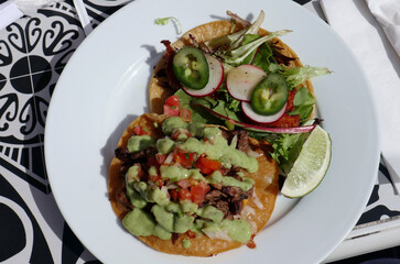 carne asada vampiro tostada and fish taco on a plate at a mexican restaurant (green avocado sauce, lime, white plate) garnished with radishes, jalapeno peppers, pico de gallo, tomatos onions cilantro