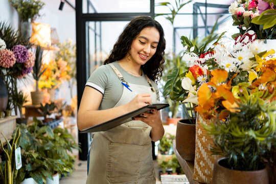 A young Latina girl with long hair works inside a flower shop dressed in an apron.The American woman is taking inventory with a notebook.Concept of working woman. Women's Day.