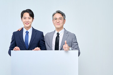 A young office worker man wearing a suit and glasses and a middle-aged man are holding a white board together and posing with various confident expressions and poses.