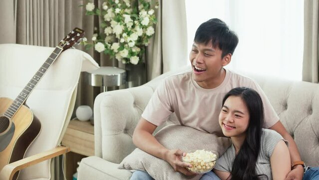 Slow motion shot of young couple sitting on sofa at home watching movie on TV and eating popcorn