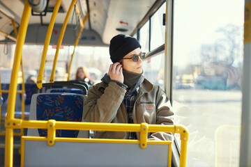 Fototapeta na wymiar Young man, passenger in stylish clothes and sunglasses sitting in modern tram and listening to music in headphones. Concept of public transport, urban lifestyle