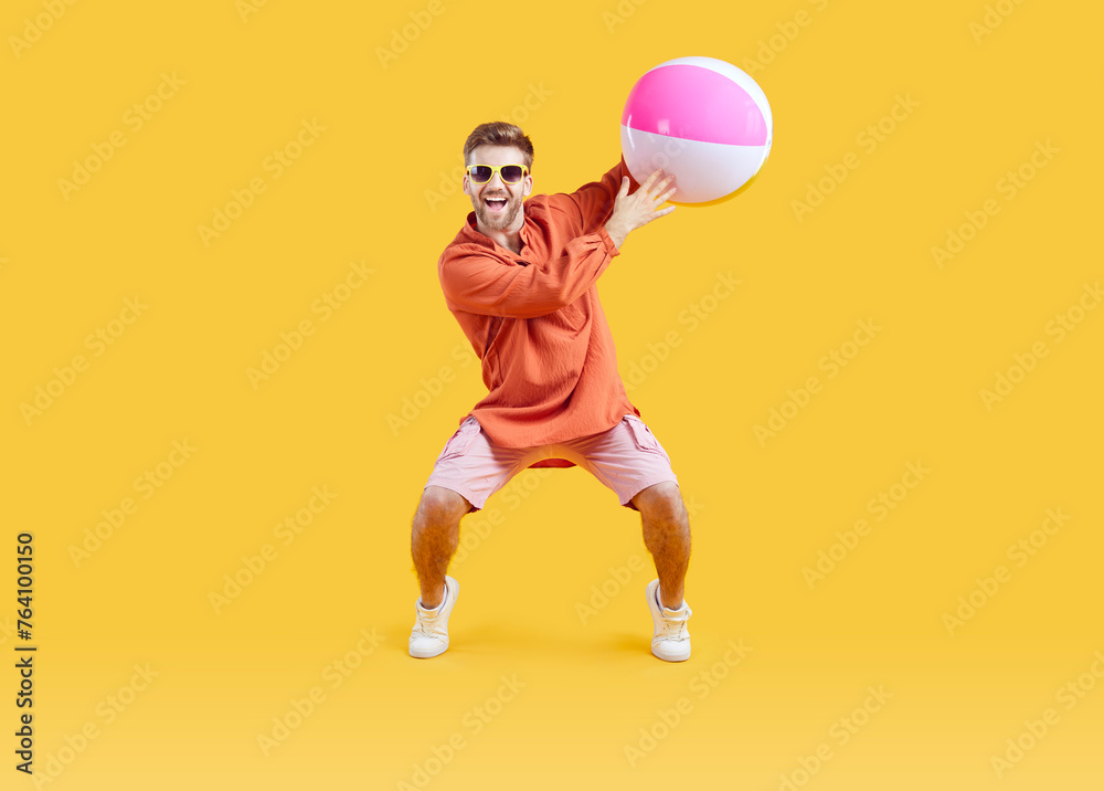 Wall mural Cheerful, funny man having fun during summer holiday. Happy handsome young guy in sun glasses, orange shirt and pink shorts playing with an inflatable beach ball isolated on a yellow color background