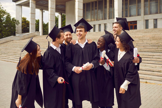 Happy diverse college or university students celebrating graduation and having fun. Group of positive multiracial multiethnic friends in black caps and gowns standing by front porch steps together