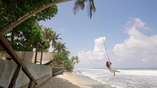 Joyful woman swings on rope over turquoise sea, white dress flowing. Holidaymaker enjoys tropical beach leisure, exhilarating freedom. Carefree moment, blue sky backdrop, palm fringed coastline. Slomo
