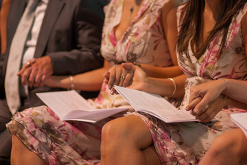 The image offers a close-up of elegantly dressed attendees seated with event programs in hand. The...