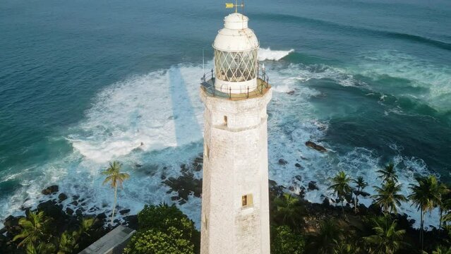 Aerial video circles historic Dondra lighthouse in Sri Lanka surrounded by greenery, white foam waves brush against rocky coast. Clear water invites travelers for exploration, relaxation.