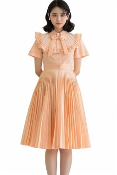 Portrait of a pretty young woman super model of Korean ethnicity showcasing a peach-colored tea dress with a cinched waist, pleated skirt, and bow accent at the neckline, styled with pearl accessories