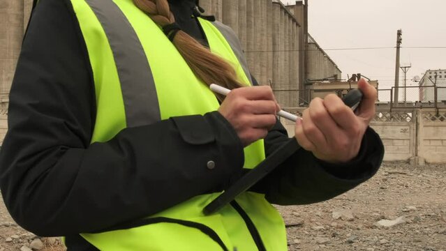 Female engineer in hard hat and reflective safety vest takes notes with stylus at building site near concrete batching plant. Engineer workers building inspection. Quality control. Slow tracking shot.