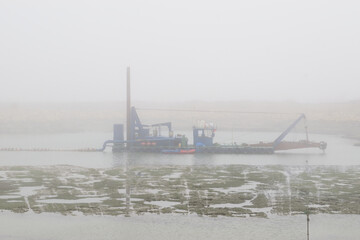 dredging vessel operating in a shallow harbor or lagoon, with muddy tidal flats in the foreground and breakwater rocks and cranes in the background. Industrial maritime scene, land reclamation