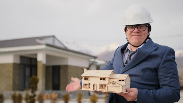 Male realtor in business suit and white helmet enthusiastically offers to buy real estate or take out mortgage, demonstrating model of future house in the suburbs.
