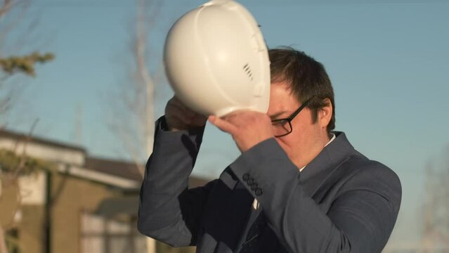 Middle-aged businessman realtor puts on protective white hardhat with a satisfied expression while adjusting his glasses while standing outdoors, slow motion.
