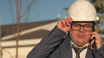 Shocked director of an enterprise in gray business suit, tie and protective white helmet adjusts his glasses talking on the phone with upset feelings. Slow motion. Close up.