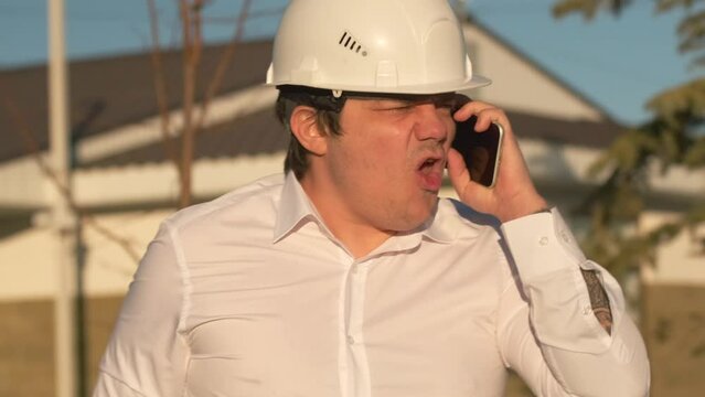 Angry project manager shouts at his interlocutor on phone, swears, commands and is angry about missed deadlines for delivery of real estate construction project.