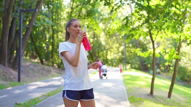 Tired young woman in sportswear drinking water from bottle and wiping sweat after jogging. Fit female athlete smiling and standing on footpath in summer park represents healthy lifestyle.