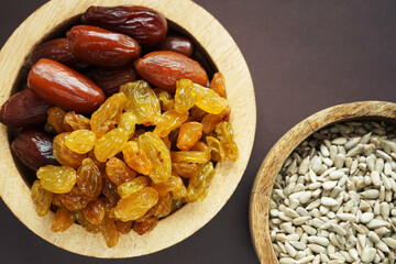 Dried dates and raisins in a wooden plate next to a mix bowl with sunflower seeds on a brown background