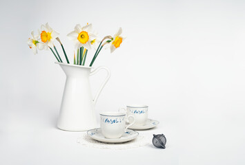 Still life with a fine porcelain tea cups and accessories arranged with a blooming bouquet of white daffodils on a textured white background - 764139182