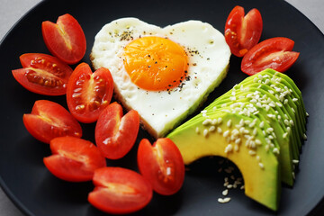 Fried egg in the shape of a heart next to sliced ​​avocado and cherry tomatoes on a black plate on a gray background