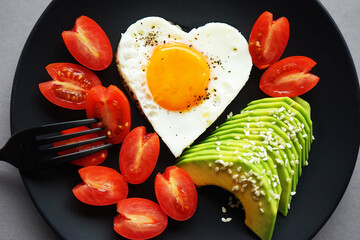 Fried egg in the shape of a heart next to sliced ​​avocado and cherry tomatoes on a black plate on a gray background